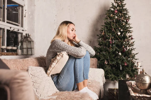 悲伤的年轻女子在圣诞前夕坐在沙发上 穿着灰色毛衣的女人 家里新年的公寓 图库图片