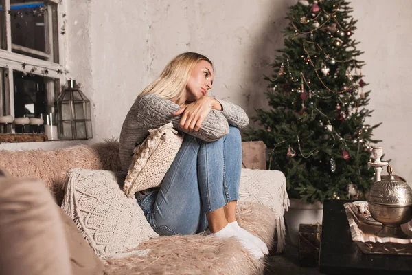 悲伤的年轻女子在圣诞前夕坐在沙发上 穿着灰色毛衣的女人 家里新年的公寓 免版税图库图片