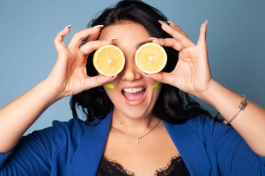 Güzel bir kız yüzüne iki tane sulu limon tutar. Lemon, ikiye böl. Siyah saçlı komik genç bir kadın stüdyoda mavi arka plandaki gözler yerine portakal dilimleri tutuyor.