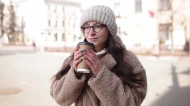 Gülümseyen Avrupalı bir kadının portresi. Şehir caddesinde geziniyor ve elinde kahveyle geziyor. Paltosu ve şapkası olan mutlu genç kadın..