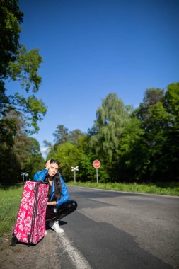 Otostop çekmekten sıkılan bir genç, güneşli bir günde yapraklı bir ormandan geçen bir yolun kenarında bekler..