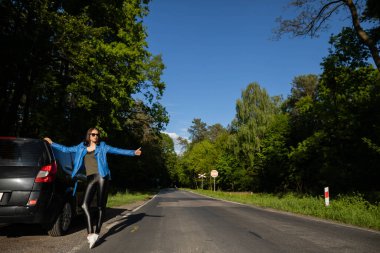 Yeşil bir ormanda yolda duran genç bir kız bozuk bir arabanın yanında yardım istiyor, diğer arabaları durduruyor..