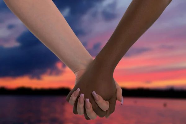 Ρομαντικό ραντεβού δύο γυναικών το ηλιοβασίλεμα δίπλα στη λίμνη. Χειραψία. Δύο γυναίκες κρατιούνται χέρι-χέρι. Αφρικανικές και ευρωπαϊκές. — Φωτογραφία Αρχείου