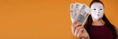 Panoramik bir çerçevede, isimsiz bir kadın elinde 100 Polonyalı zloti banknotuyla duruyor ve çiziyor. Yüzü yok çünkü üzerinde maske var..