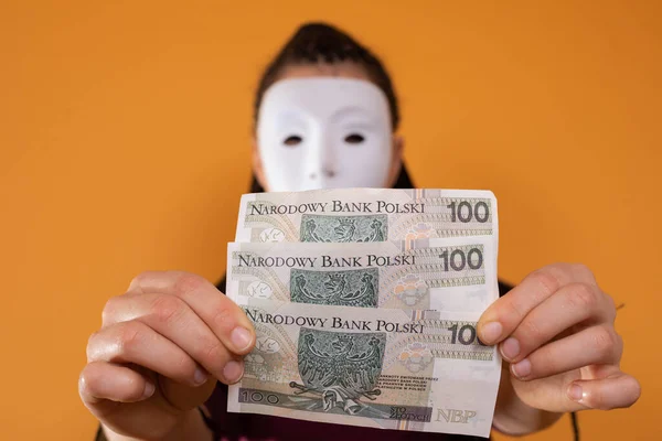 An anonymous woman stands and pulls her hand in with paper banknotes with the face value of one hundred Polish zlotys. No face because theres a mask on it.