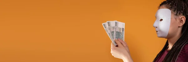 In a panoramic frame, an anonymous woman stands and draws in her hand with paper banknotes of one hundred Polish zlotys. No face because theres a mask on it.