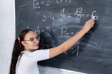 Genç bir kız, beyaz tebeşirle okul tahtasına yazı yazarak okuldaki matematik problemini çözüyor. Saçları iki at kuyruklu pembe bir kurdeleyle tutturulmuş..