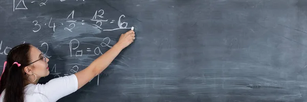 Panoramisch schot. Een tienermeisje lost een wiskundeprobleem op school op door op een schoolbord met wit krijt te schrijven. Haar haar is gepind met een roze lint in twee paardenstaarten. — Stockfoto