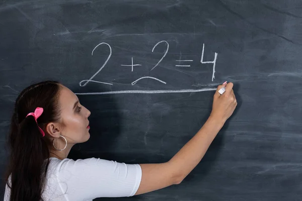 Tiener krijt een wiskundige vergelijking op een donkergrijs schoolbord. Haar haar is gepind met een roze lint in twee paardenstaarten. — Stockfoto