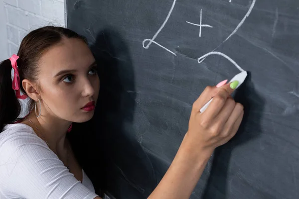 Tiener krijt een wiskundige vergelijking op een donkergrijs schoolbord. Haar haar is gepind met een roze lint in twee paardenstaarten. — Stockfoto