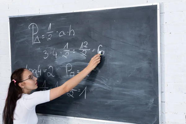 Teini-ikäinen tyttö ratkaisee koulussa matematiikan ongelmaa kirjoittamalla koulun liitutaululle valkoisella liidulla. Hänen hiuksensa on kiinnitetty vaaleanpunaisella nauhalla kahdessa poninhännässä. — kuvapankkivalokuva