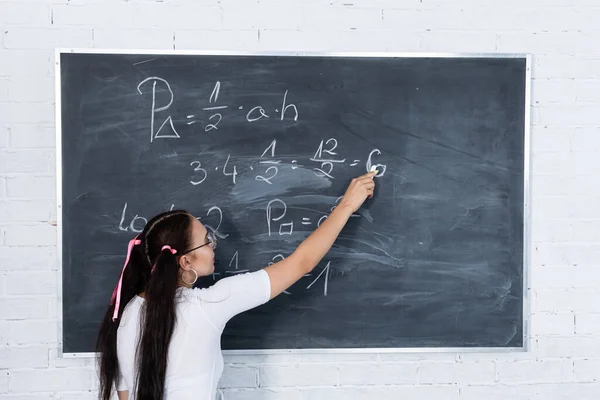 Genç bir kız, beyaz tebeşirle okul tahtasına yazı yazarak okuldaki matematik problemini çözüyor. Saçları iki at kuyruklu pembe bir kurdeleyle tutturulmuş.. — Stok fotoğraf