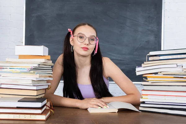 Ein Teenager sitzt im Klassenzimmer zwischen Bücherstapeln und beginnt, die erste Lektüre zu lesen. Der Schüler beginnt zu lernen. Ihr Haar ist mit einer rosafarbenen Schleife in zwei Pferdeschwänze gesteckt. — Stockfoto