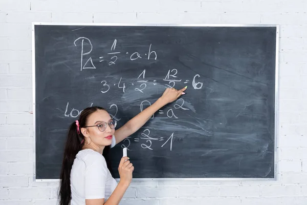 Genç bir kız, beyaz tebeşirle okul tahtasına yazı yazarak okuldaki matematik problemini çözüyor. Saçları iki at kuyruklu pembe bir kurdeleyle tutturulmuş.. — Stok fotoğraf
