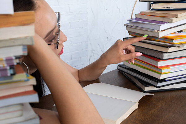 Студентка ищет следующую школьную книгу, чтобы прочитать среди всех масс, которые находятся на столе рядом с ней. Её волосы прижаты розовой лентой в двух хвостах..