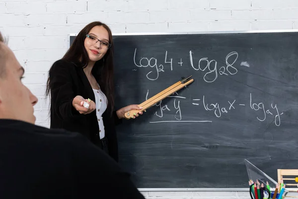 Een wiskundeleraar nodigt haar leerling uit, geeft hem een krijt, naar het schoolbord zodat hij het probleem met logaritmen kan blijven oplossen.. — Stockfoto