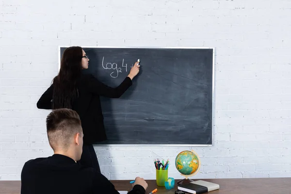 Молодий вчитель математики пише на дошці з крейдою. Довге чорне волосся. Освіта в середній школі та коледжі . — стокове фото
