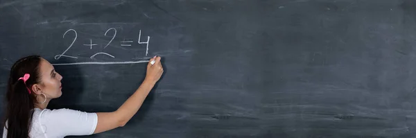 Panoramisch uitzicht als tienermeisje krijt een wiskundige vergelijking op een donkergrijs schoolbord. Haar haar is gepind met een roze lint in twee paardenstaarten. — Stockfoto