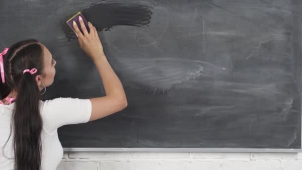 Stávající teenager utře houbičkou tabuli. Školní tabule visí na bílé cihlové zdi. Pomocná dívka stojí na tabuli. — Stock video