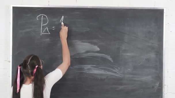 Teenager kreidet eine mathematische Gleichung auf eine dunkelgraue Schultafel. Ihr Haar ist mit einer rosafarbenen Schleife in zwei Pferdeschwänze gesteckt. — Stockvideo
