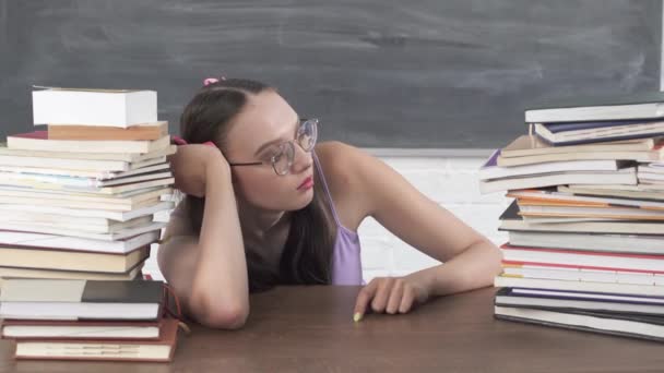 Una adolescente se sienta en el aula entre una pila de libros y comienza a buscar su primera lectura. El estudiante comienza a aprender. Su cabello está cubierto con una cinta rosa en dos colas de caballo. — Vídeo de stock