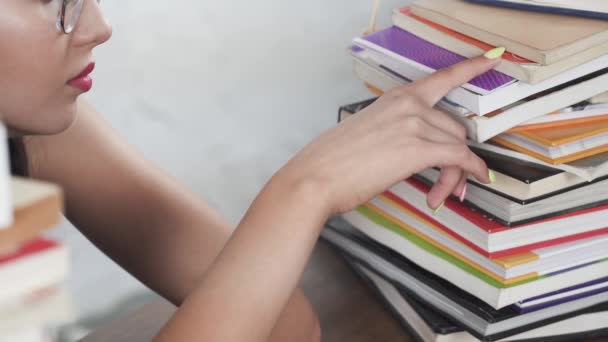 Un'adolescente si siede in classe tra un mucchio di libri e inizia a cercare la sua prima lettura. Lo studente inizia a imparare. I suoi capelli sono appuntati con un nastro rosa in due code di cavallo. — Video Stock