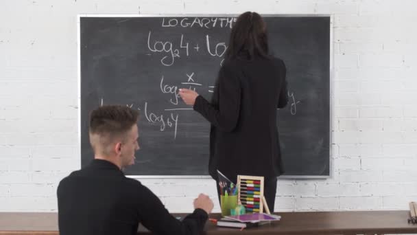 Educação no ensino médio e na faculdade. O jovem professor de matemática escreve num quadro negro com giz. Longo cabelo preto solto. — Vídeo de Stock