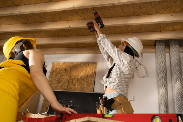 La ingeniera le muestra a su colega del sitio de construcción cómo atornillar los tornillos en las vigas por encima de su cabeza con un destornillador. — Foto de Stock