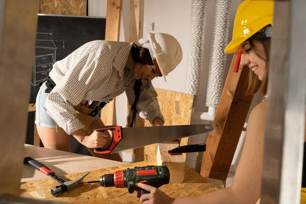 Una morena corta un tronco de madera con una sierra de mano. Chica sonriente mientras trabaja en un sitio de construcción. — Foto de Stock