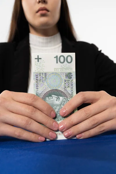 Una joven morena sostiene delante de ella una pila de billetes con un valor facial de cien zlotys polacos. Una chica está sentada en una silla a la mesa con un mantel azul. — Foto de Stock