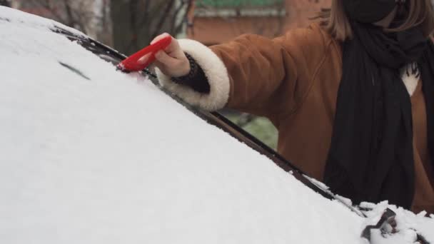 Una giovane ragazza sta spalando la neve dal parabrezza dell'auto con uno speciale triangolo rosso di plastica. La prima neve a Lublino. — Video Stock