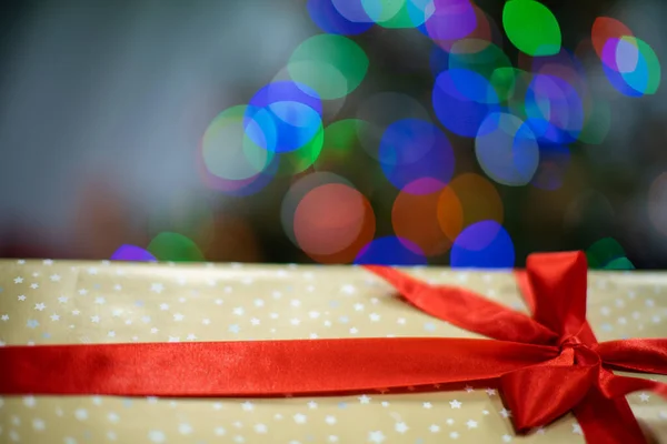 Atado con una cinta roja y un lazo grande. Caja de regalo con lazo. Un regalo en Navidad. Luces brillantes del árbol de Navidad en el fondo. —  Fotos de Stock