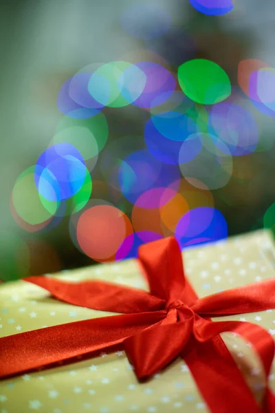 Atado con una cinta roja y un lazo grande. Caja de regalo con lazo. Un regalo en Navidad. Luces brillantes del árbol de Navidad en el fondo. —  Fotos de Stock