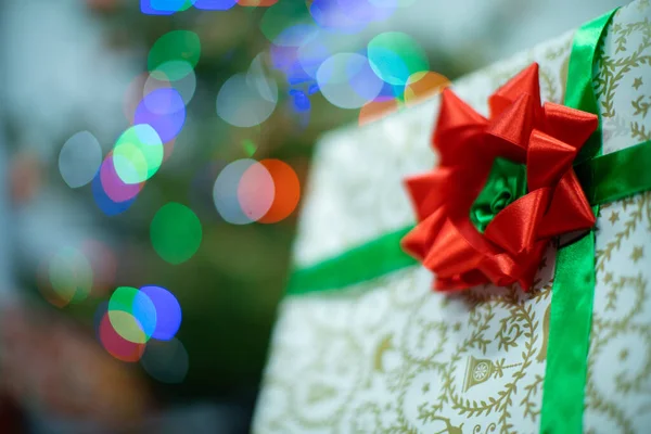 Un regalo atado con una cinta verde con un lazo rojo. Caja de regalo con lazo. Un regalo en Navidad. Luces brillantes del árbol de Navidad en el fondo. — Foto de Stock