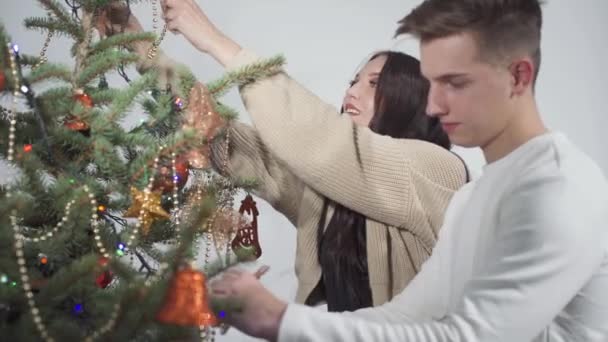 Mladý pár zdobí vánoční stromek k Vánocům. Dívka a chlapec zdobí smrk se zlatými vánočními míčky a lesklými řetězy. — Stock video