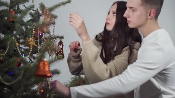 Una joven pareja decora un árbol de Navidad para Navidad. Una niña y un niño decoran el abeto con bolas de Navidad doradas y cadenas brillantes. — Vídeos de Stock