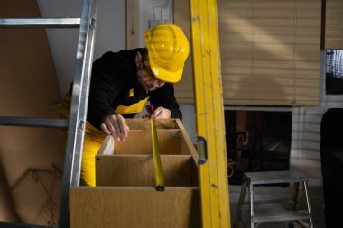 Bir inşaat işçisi üst kattaki dolabı mezurayla kontrol ediyor. Kişisel koruyucu ekipman giyiyordu. Deneyimli inşaat işçisi.