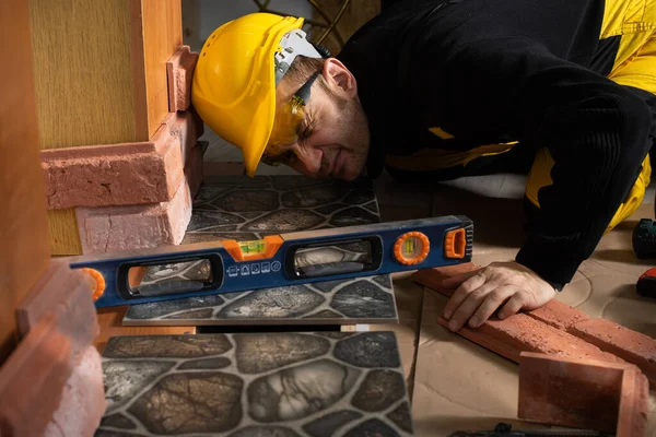 Un trabajador de la construcción comprueba con un nivel de espíritu que las baldosas de terracota se colocan uniformemente en todas las áreas. Vestido con equipo de protección personal. — Foto de Stock