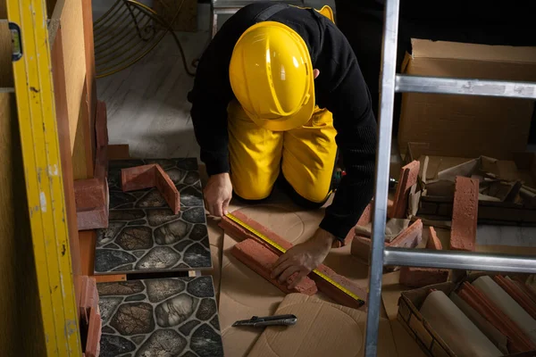 A construction worker in working clothes, kneeling, measures the retractable decorative tiles in the shape of old red brick with a tape measure. - Stock-foto