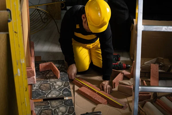 A construction worker in working clothes, kneeling, measures the retractable decorative tiles in the shape of old red brick with a tape measure. — Stockfoto