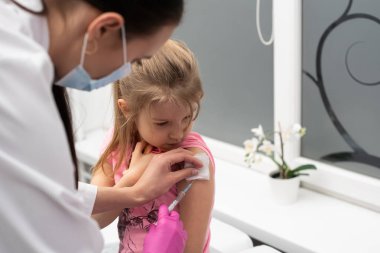 Doktor küçük bir kıza enjeksiyon yapıyor. Çocuk boğma kabına bakıyor. Küçük çocuklar için önleyici aşı.