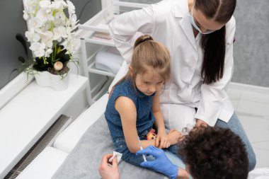 Doktor küçük bir kızı aşılamaya hazırlanıyor. Çocuğu güvende hissettirmek için genç bir hemşire çocuğun yanına oturur. Küçük çocuklar için önleyici aşı.
