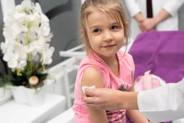 La chica está mirando fuera del marco. Una joven doctora está preparando a un niño para una inyección desinfectando un pedazo de piel en el brazo con gasa y productos médicos. Vacuna preventiva para — Foto de Stock