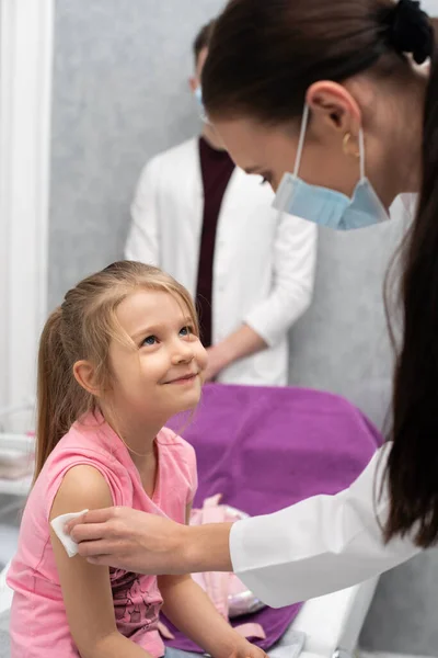 La niña mira a la enfermera con una sonrisa y confianza. Una joven doctora está preparando a un niño para una inyección desinfectando un pedazo de piel en el brazo con gasa y productos médicos — Foto de Stock