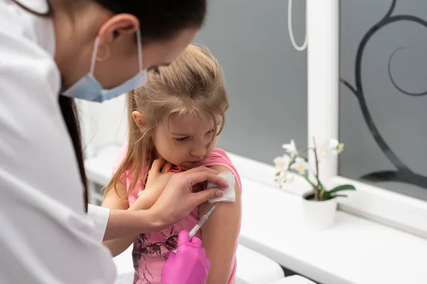 El médico le va a poner una inyección a una niña. El niño mira fijamente en un tazón de estrangulamiento. Vacuna preventiva para niños pequeños. — Foto de Stock