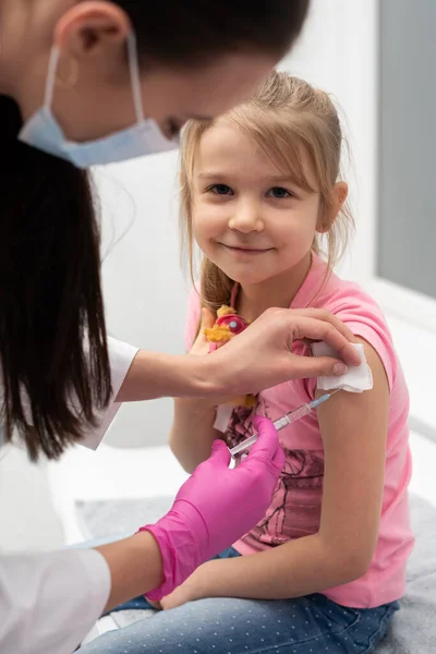 Mientras recibe la vacuna, la niña sonriente mira directamente a la lente. El médico apuñala al niño en el brazo con una aguja. Vacuna preventiva para niños pequeños. — Foto de Stock