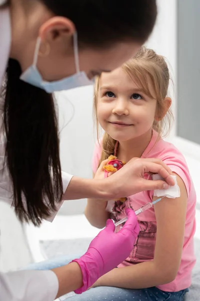 Mientras recibe la vacuna, la niña sonríe suavemente y mira con confianza a los ojos de las enfermeras. El médico apuñala al niño en el brazo con una aguja. Vacuna preventiva para niños pequeños. — Foto de Stock