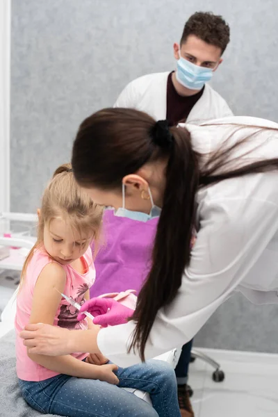 El médico le va a poner una inyección a una niña. El niño mira fijamente en un tazón de estrangulamiento. Vacuna preventiva para niños pequeños. Vista de cerca. El médico está parado atrás.. — Foto de Stock