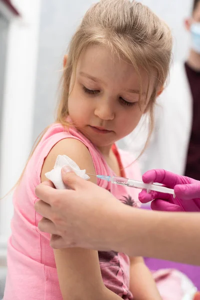 El médico le va a poner una inyección a una niña. El niño mira fijamente en un tazón de estrangulamiento. Vacuna preventiva para niños pequeños. Vista de cerca. El médico está parado atrás.. — Foto de Stock