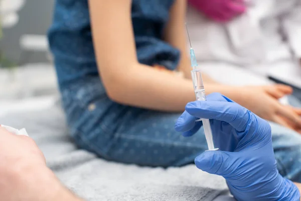 El médico sostiene una jeringa con una aguja en la que se encuentra la vacuna. En el fondo se puede ver que un niño pequeño la está esperando. Vacuna preventiva para niños pequeños. Primer plano — Foto de Stock